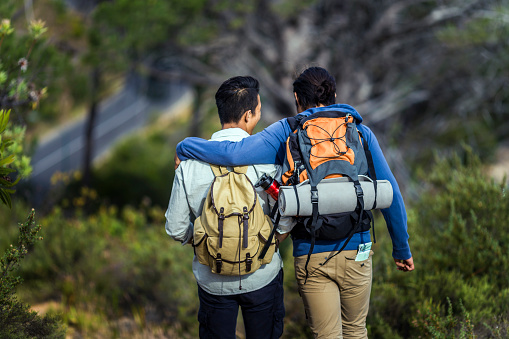 A photo of young gay hikers walking in forest. Rear view of travellers carrying backpacks in woodland. Young man is walking arm around boyfriend during vacation.