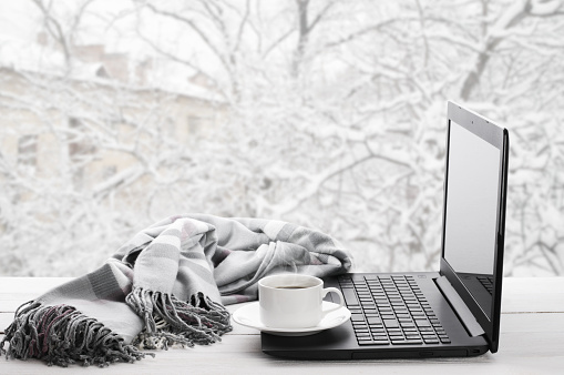 Cozy winter still life: laptop, cup of hot coffee and warm plaid on windowsill against snow landscape from outside.