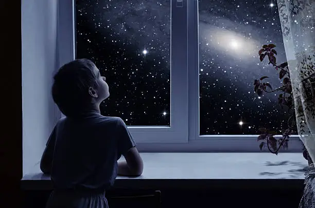 A little boy is standing near the window and looking outside, imagining boundless space with myriad of stars