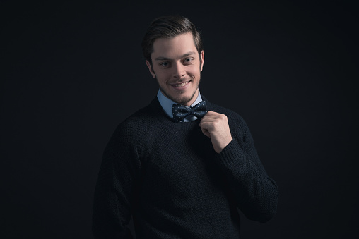 Smiling man wearing dark blue sweater and light blue shirt holding bow tie.