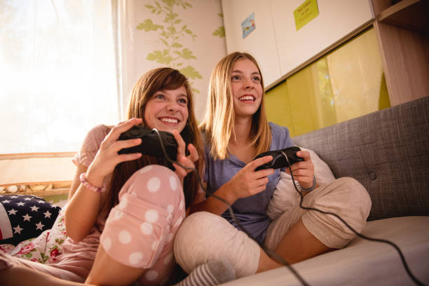 Smiling adolescent girl friends playing competitive computer games on a bed next to each other at a daytime pyjama party