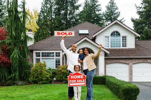 Portrait of a young family standing in front of house, holding a Home For Sale sign with arms raised enthusiastically.