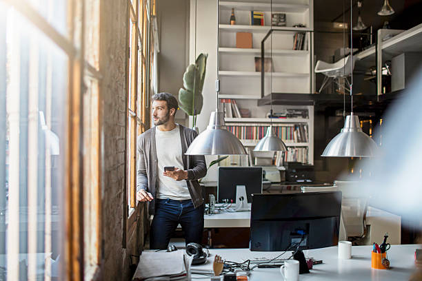 Young entrepreneur looking through the window in the office Young entrepreneur looking through the window in the office on a sunny day, he's holding the mobile phone waiting for an important call - BOOK COVERS IN THE BACKGROUND HAS BEEN MODIFIED. stars in your eyes stock pictures, royalty-free photos & images