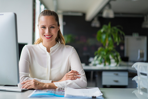 A photo of beautiful businesswoman sitting at desk. Portrait of happy female professional is in office. Smiling executive is in formals at workplace.