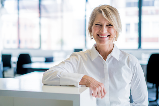 A photo of happy businesswoman leaning on cubicle in office. Portrait of smiling female professional in formals. Executive is at brightly lit workplace.