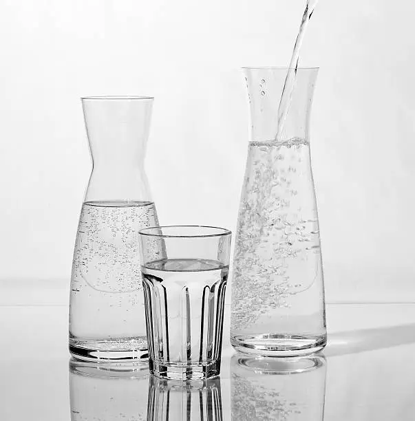 Two carafes and a drinking glass are standing on a table. Water is filled in a carafe, the other carafe and the drinking glass are already filled with drinking water.