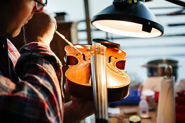 Close up shot of a violin maker repairing an old valuable violin in his workshop. The workspace is illuminated by artificial light.