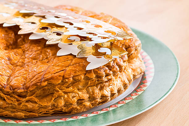 Horizontal composition color photography of close-up in selective focus of homemade Epiphany King's cake pastry with marzipan (frangipane), called "Galette des Rois" in french in his plate with a gold paper crown placed on top to celebrate Epiphany. A small porcelain figurine is hidden inside the cake, and the person who finds it becomes king or queen.