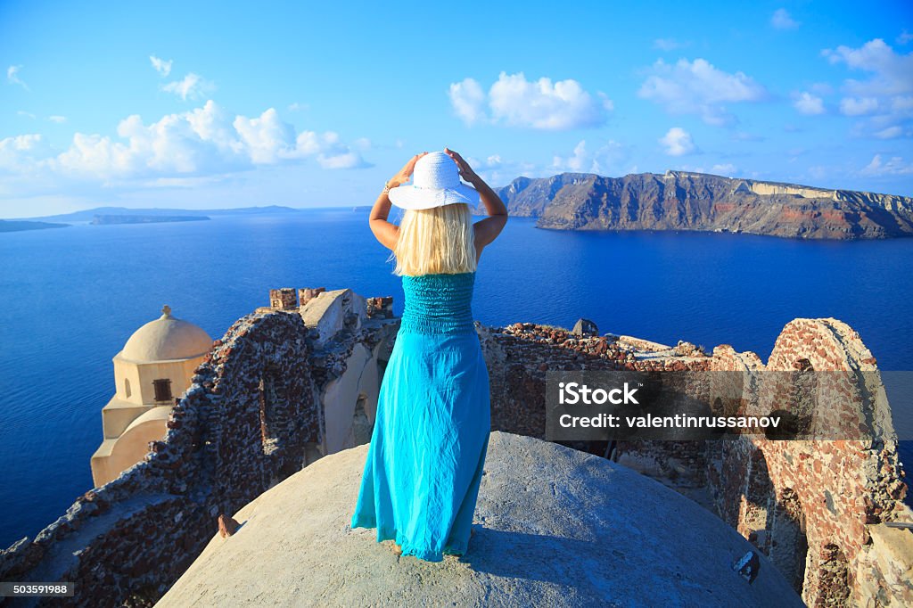 Woman with turquoise dress in Santorini, Greece. Rear view of beautiful blond woman in a turqoise dress standing over the rooftops in Oia, Santorini, Greece. Who admires the beauty of the volcano and the sea. With hands holding the head and wear beach hat. Fun Stock Photo