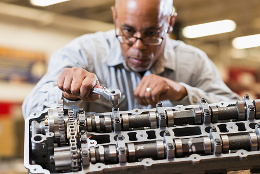 A mature, African American man in an auto repair shop working on a gasoline engine.  He is wearing a gray shirt, looking down at the engine with a serious expression.  The focus is on his hand and the tool he is using.