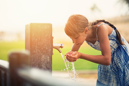 Little girl aged 9 drinking water from a public drinking fountain. Sunny summer day. The girl is wearing blue sun dress.