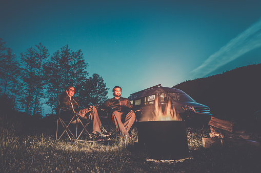 Heterosexual Couple Sitting Next to the Campfire with a Camper Van in the background. They are holding their hands.