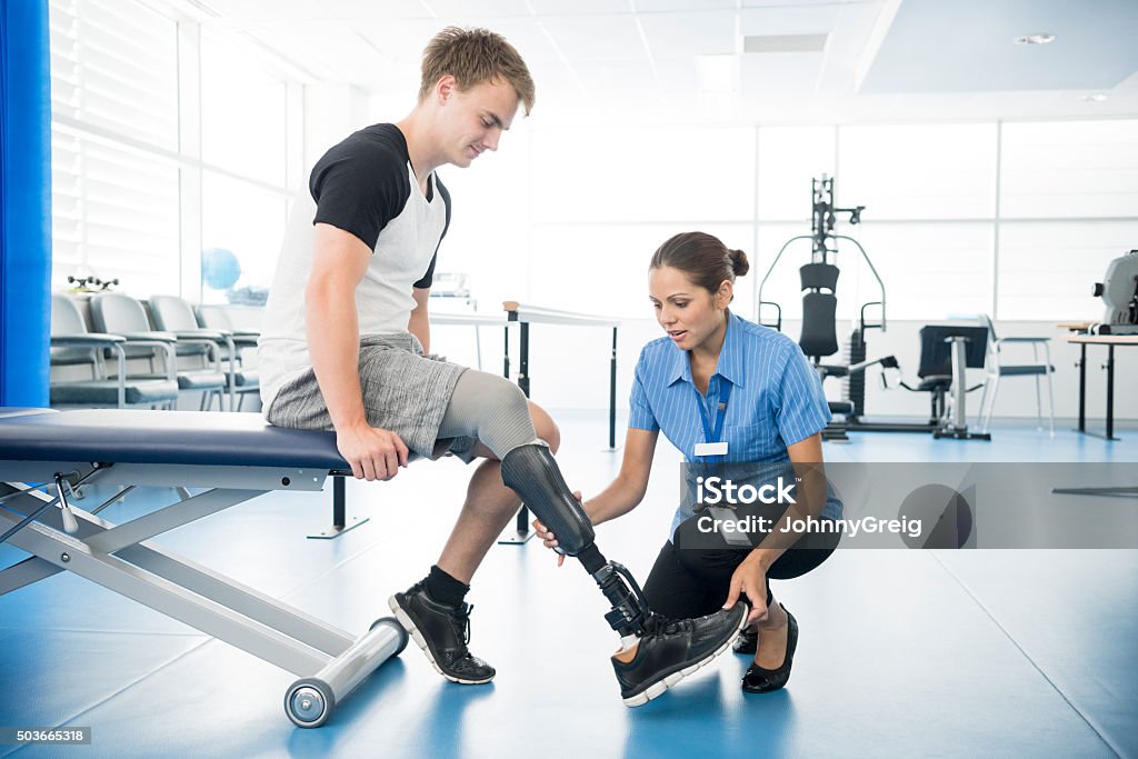 Female physiotherapist helping young man with prosthetic leg Patient sitting on bed with nurse holding prosthetic limb. Male amputee with prosthesis in hospital. Prosthetic Equipment Stock Photo