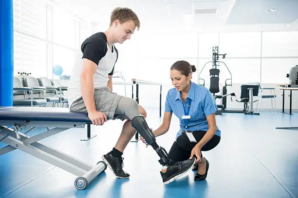 Patient sitting on bed with nurse holding prosthetic limb. Male amputee with prosthesis in hospital.