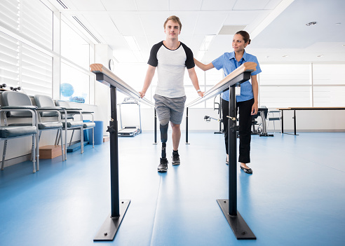 Female physiotherapy helping young male amputee to walk. Patient wearing prosthesis using rehabilitation equipment. Man learning to walk again. Rehabilitation, recovery, determination, physiotherapy.