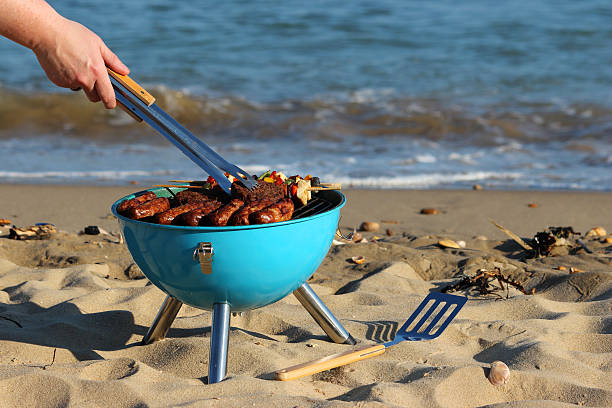 Image of beach barbecue, seaside BBQ, sausages, burgers, kebabs Photo showing a beach barbecue on the sands of a popular beachfront resort, with the gentle waves of the blue sea lapping on the shore in the background of the picture.  This seaside barbecue is pictured with freshly cooked sausages, beef burgers and chicken shish kebabs on bamboo skewers (bell peppers, red onions and pieces of chicken breast) on the stainless steel grill, above the hot coals, while a spatula is placed on the sand, ready to flip / serve up the burgers. coke coal stock pictures, royalty-free photos & images