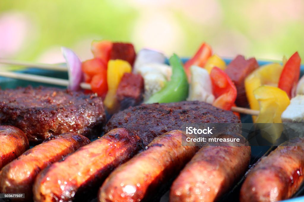 Kettle charcoal barbecue BBQ in garden, sausages, burgers, kebabs image Photo showing a portable kettle barbecue / charcoal BBQ in a garden setting, stood on a wooden patio table with the lawn and pink hardy geraniums forming the blurred background.  Pictured cooking on the barbecue are some sausages, beef burgers and colourful kebabs, made from red, green and yellow bell peppers, red onions and chicken pieces. Barbecue - Meal Stock Photo