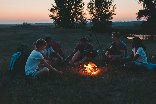 Friends camping together in nature. Sitting on grass near campfire, singing and playing acoustic guitar. Fist pot is on fire. Three mails and two females. Sunset in background. Summer or spring time.Deliblatska pescara, Bela Crkva, Serbia, Europe