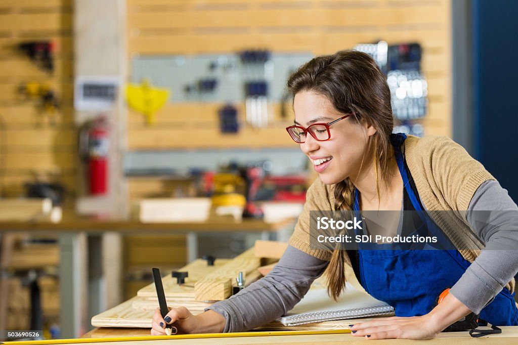 Young artist smiling while creating something with wood in makerspace Young adult Caucasian woman is measuring boards while working in carpentry shop or makerspace. Young artist is smiling while using a measuring tape to measure boards. Work benches and tools are organized in background. Accuracy Stock Photo