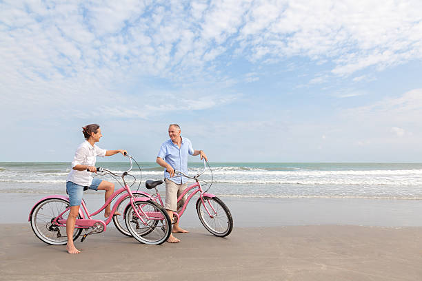 matura coppia biciclette equitazione all'aperto sulla spiaggia al tramonto - mature adult bicycle senior adult heterosexual couple foto e immagini stock