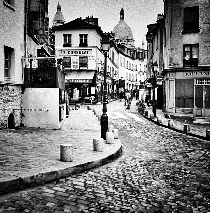 Paris, France - May 9, 2015: These are black and white, monochrome images of the Montmartre district of Paris. This is known as the artist area of Paris and is a famous tourist destination. It is an overcast afternoon in Spring, March. This is a corner with cobbled streets and old buildings. There are people in the distance.
