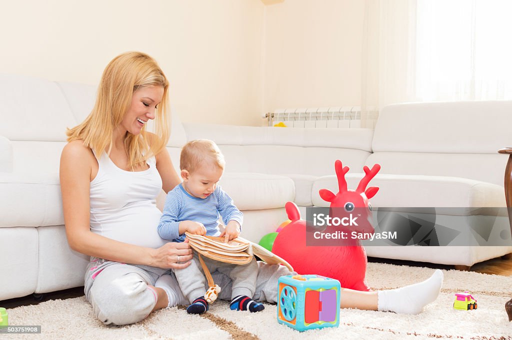 Curious boy playing with his pregnant mother Pregnant mother and her son are playing in living room with a toys. They are having a great time together. Mother is reading a book to her son. 12-23 Months Stock Photo