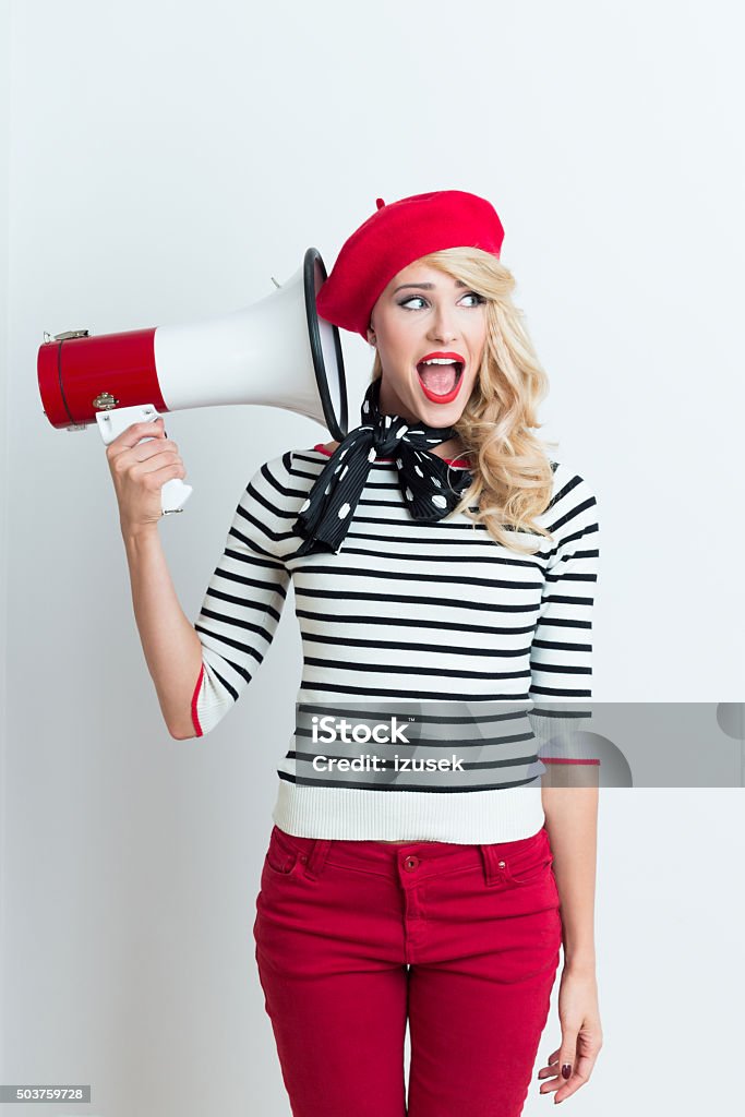 Blonde french woman wearing red beret holding a megaphone Portrait of excited beautiful blonde woman in french outfit, wearing a red beret, striped blouse and neckerchief, holding a megaphone next to her ear, looking away with mouth open. Adult Stock Photo