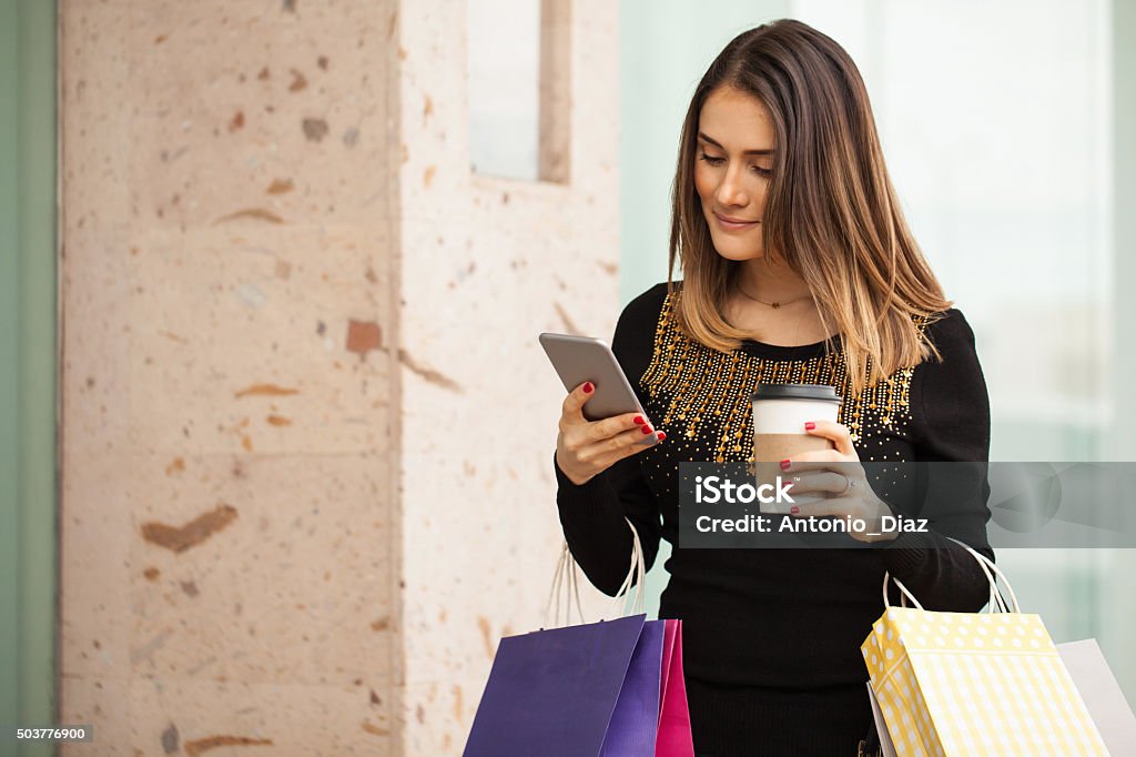 Cute woman using a phone at the shopping mall Beautiful brunette using her smartphone and drinking coffee while doing some shopping at a mall 20-29 Years Stock Photo