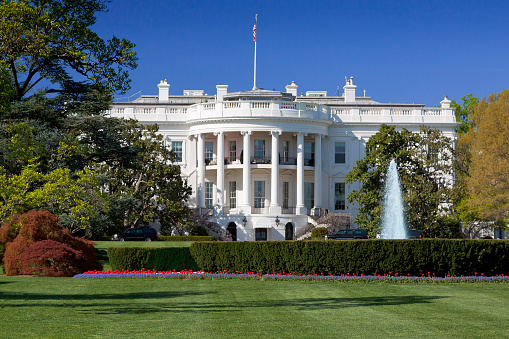 The South Portico of the White House. Washington DC. The White House is the official residence and principal workplace of the President of the United States, located at 1600 Pennsylvania Avenue. Beautifully landscaped lawn with flowers, fountain and blooming trees is in foreground. Deep blue clear sky is in background. American flag is flying atop. The image lit by spring evening sun. Canon 24-105mm f/4L lens.