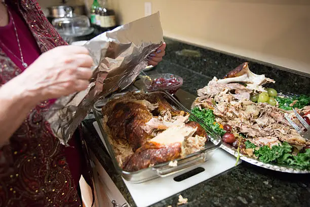 Caucasian woman whose face isn't visible is covering a glass pan of turkey leftovers with tin foil. The platter the turkey was on is beside it with the carcass and bits of turkey on it. They are sitting on a granite kitchen counter. Taken with a Canon 5D Mark 3.