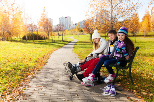 Two girls and a boy putting on roller blades sitting on the bench in the park