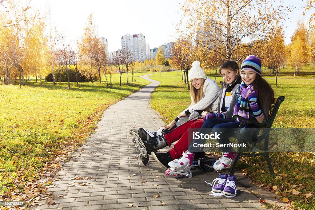 Three kids getting ready to skate Two girls and a boy putting on roller blades sitting on the bench in the park Autumn Stock Photo