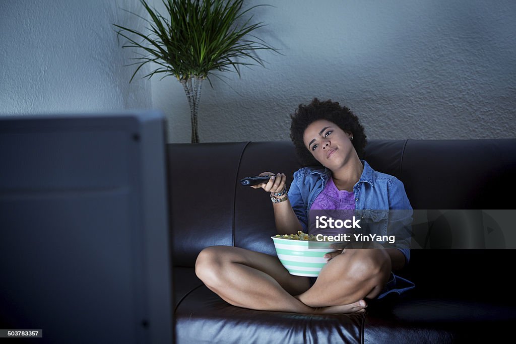 Young Audience Bored To Death Surfing TV Programming_ A bored couch potato with remote control surfing and watching TV on the couch alone with popcorn. Television Set Stock Photo