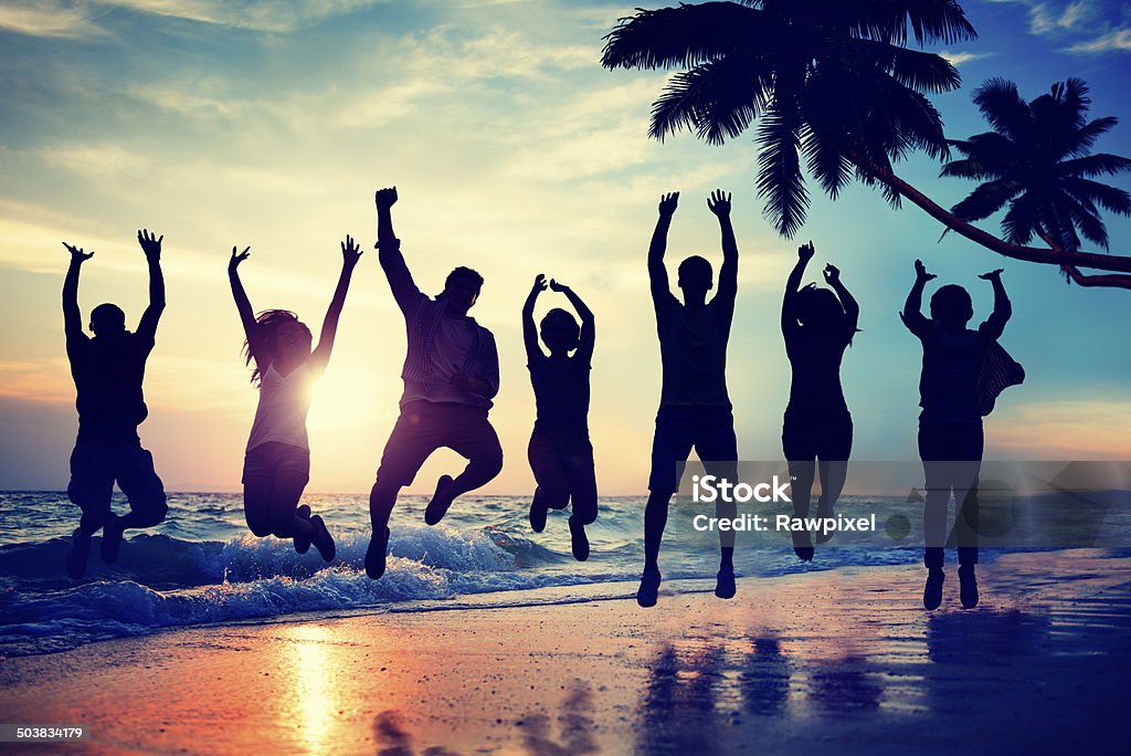 Group jumping on beach at sunset Young people are shown jumping excitedly in this image.  They are on a beach as the sun is setting.  The people are shown as silhouettes.  There are seven young people.  Surf is washing in onto the beach just under their legs. In the right of the image is a palm tree leaning over the beach, as well three young people whose shadows reflect off the water from the surf just below them.  The sun is setting above the water between the people, creating an orange line in the sky's horizon. Multiracial Group Stock Photo