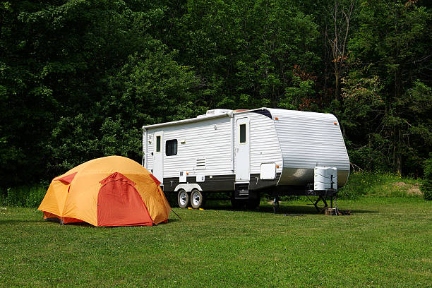 Summer camping image of a medium sized travel trailer parked alongside a bright orange dome tent on a wooded campsite..
