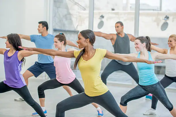A multi-ethnic group of young adults are taking a yoga class together and are holding warrior two pose.