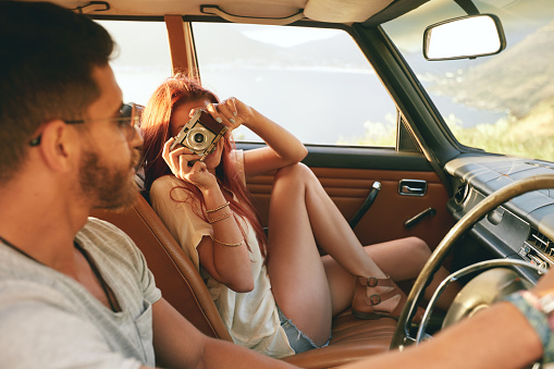 Caucasian couple going on a road trip. Woman taking pictures with camera and man driving car.