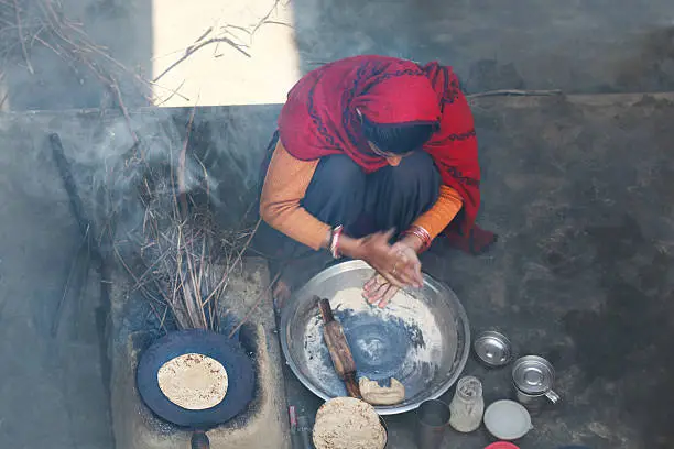 Indian Women Sitting in Rural Environment wearing Suit which is Traditional Dress for Women In Rural India & Making chapatti at Home on wood Burning Stove. There is Mud stove with continuous wood fire. 