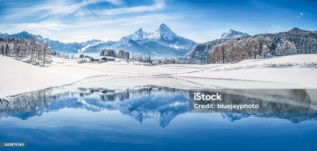 Winter wonderland in the Alps reflecting in crystal-clear mountain lake Panoramic view of beautiful white winter wonderland scenery in the Alps with snowy mountain summits reflecting in crystal clear mountain lake on a cold sunny day with blue sky and clouds. Winter Stock Photo