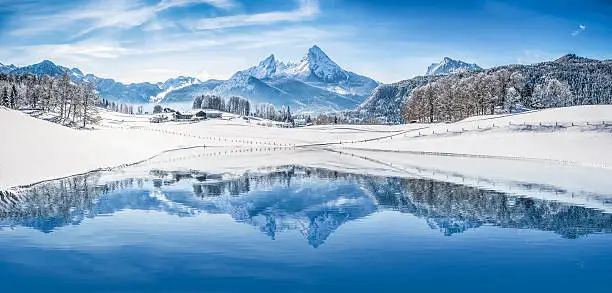 Panoramic view of beautiful white winter wonderland scenery in the Alps with snowy mountain summits reflecting in crystal clear mountain lake on a cold sunny day with blue sky and clouds.