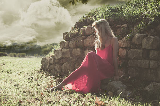fairy like young woman in red dress and wreath of flowers in hair sit by old wall in nature, full body shot, side view
