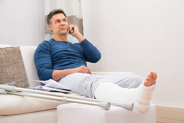 Disabled Man Talking On Cellphone Man With Fractured Leg Sitting On Sofa Talking On Cellphone broken leg stock pictures, royalty-free photos & images