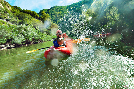 A male is looking back at camera and splahsing in red inflatable canoe having fun in calm waters of a river.