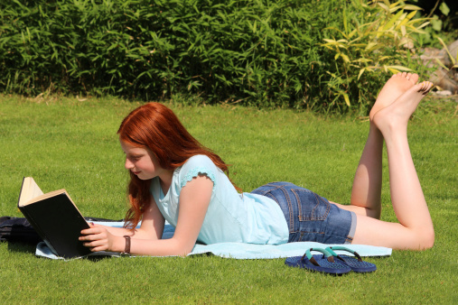 Photo showing a young girl reading her school book outside on the neatly mown garden lawn, lying in the sunshine on a towel, so that she can sunbathe whilst studying.