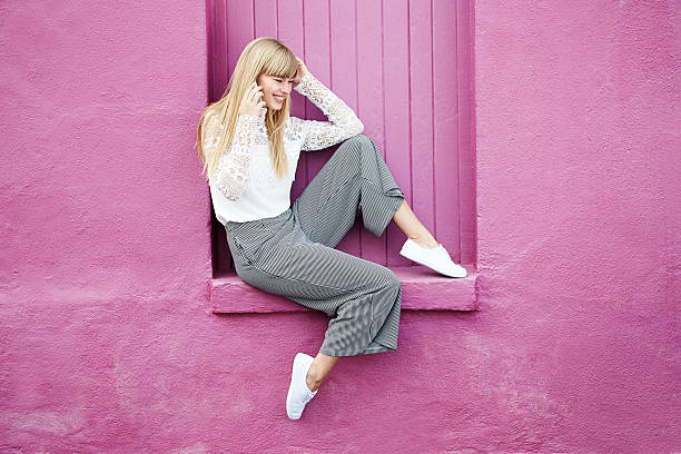 Young woman on the phone Young woman on the phone sitting on pink wall cropped pants photos stock pictures, royalty-free photos & images