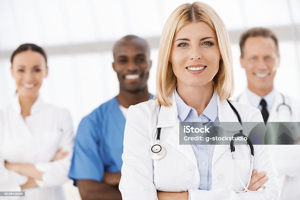 Confident in her team. Confident in her team. Beautiful female doctor keeping arms crossed and smiling while her colleagues standing behind her in the background Female Doctor Stock Photo