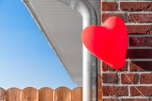 Part of a brick build house with water drain pipe and board fence and a floating red heart shaped balloon at the corner as a dream of owning a house