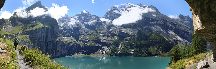 Kandersteg, Switzerland - August 13, 2008: in the Swiss Alps at 1500 m elevation lies the Oeschinen lake, surrounded by high snowcapped peaks; two hikers stroll on a foothpath high above the lake