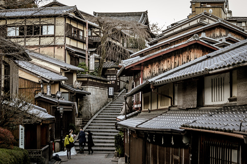Kyoto, Japan - November 27, 2015: Tourists walk along a narrow road too much traveled, Kiyomizu Zaka street is an ascending narrow lane with pretty shops selling foods, souvenirs, clothing, arts and small shrines. As you walk along this alley, it feels like jumping into a different era, an alternate history of a mixture of modern and traditional Japan, that is different from Gion. Flows of crowds going up and down the street, kids in student uniforms, geishas, older people in overcoats, teenagers, blend together. The shops look unique in each own way. Before you realize, the street ended at the giant Deva gate of Kiyomizudera.