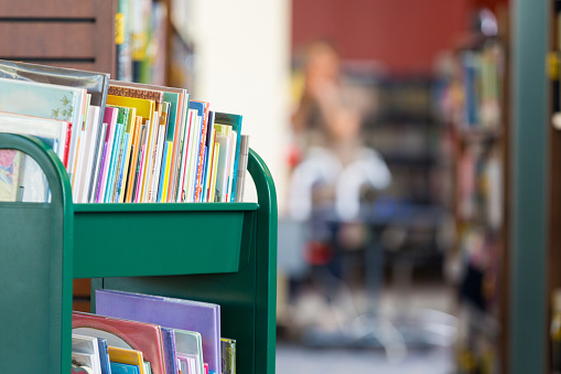 Green rolling cart full of colorful childrens books sitting in front of large bookshelves in modern public library or elementary school library.